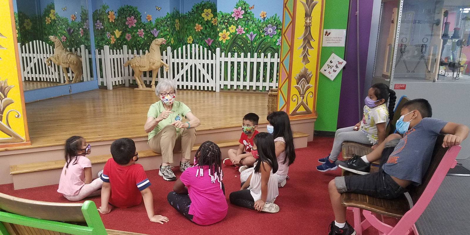 Woman with blonde hair and green polo shirt reading a story to young children gathered around and sitting on plastic tree stumps