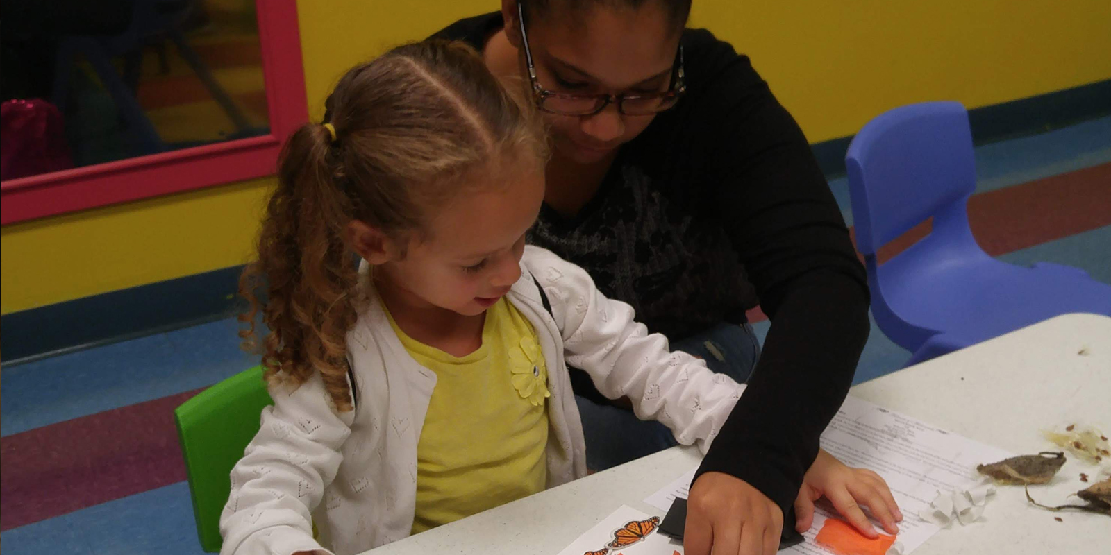 Young woman wearing dark-rimmed glasses helping young girl with light brown hair and pigtails with a paper craft