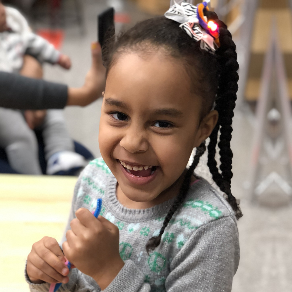 Young girl with dark braided hair and gray sweater smiling holding pipe cleaners
