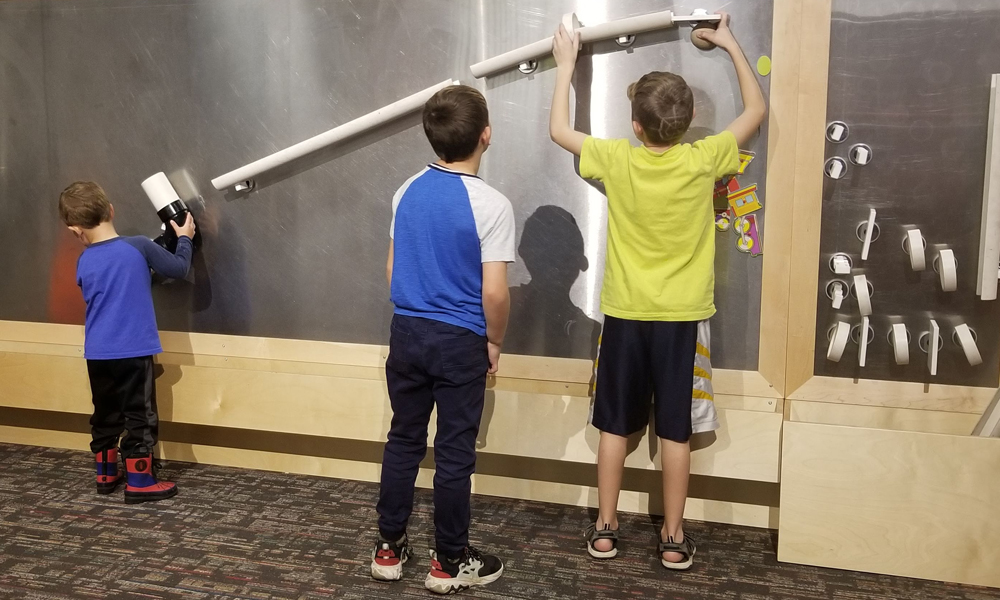 Three young children playing with a tube sloping down and attached to a wall