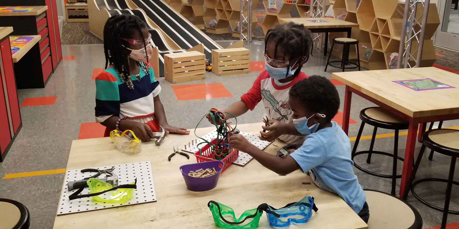 Young children wearing masks and goggles at a table with tools and wooden pegboard