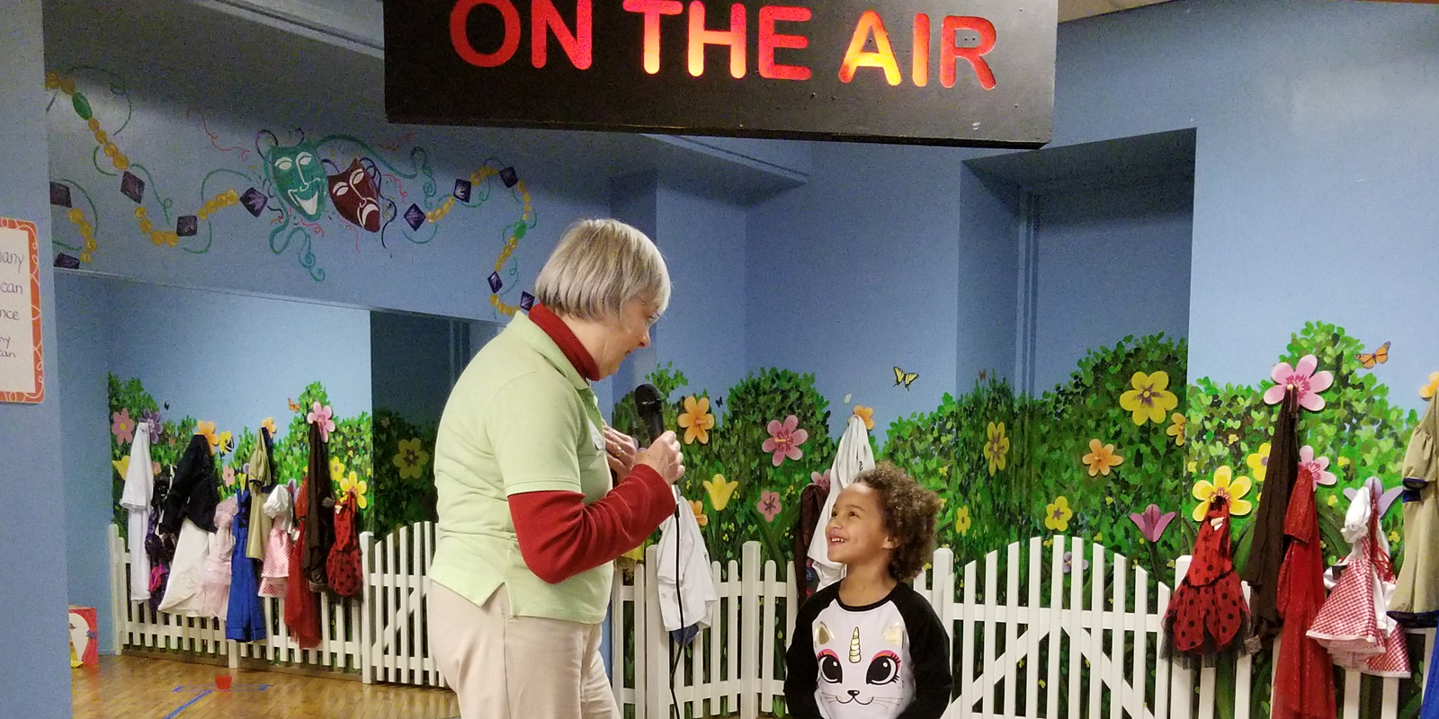Woman with blonde hair and green polo shirt and khakis standing next to young girl under a sign that reads On the Air.