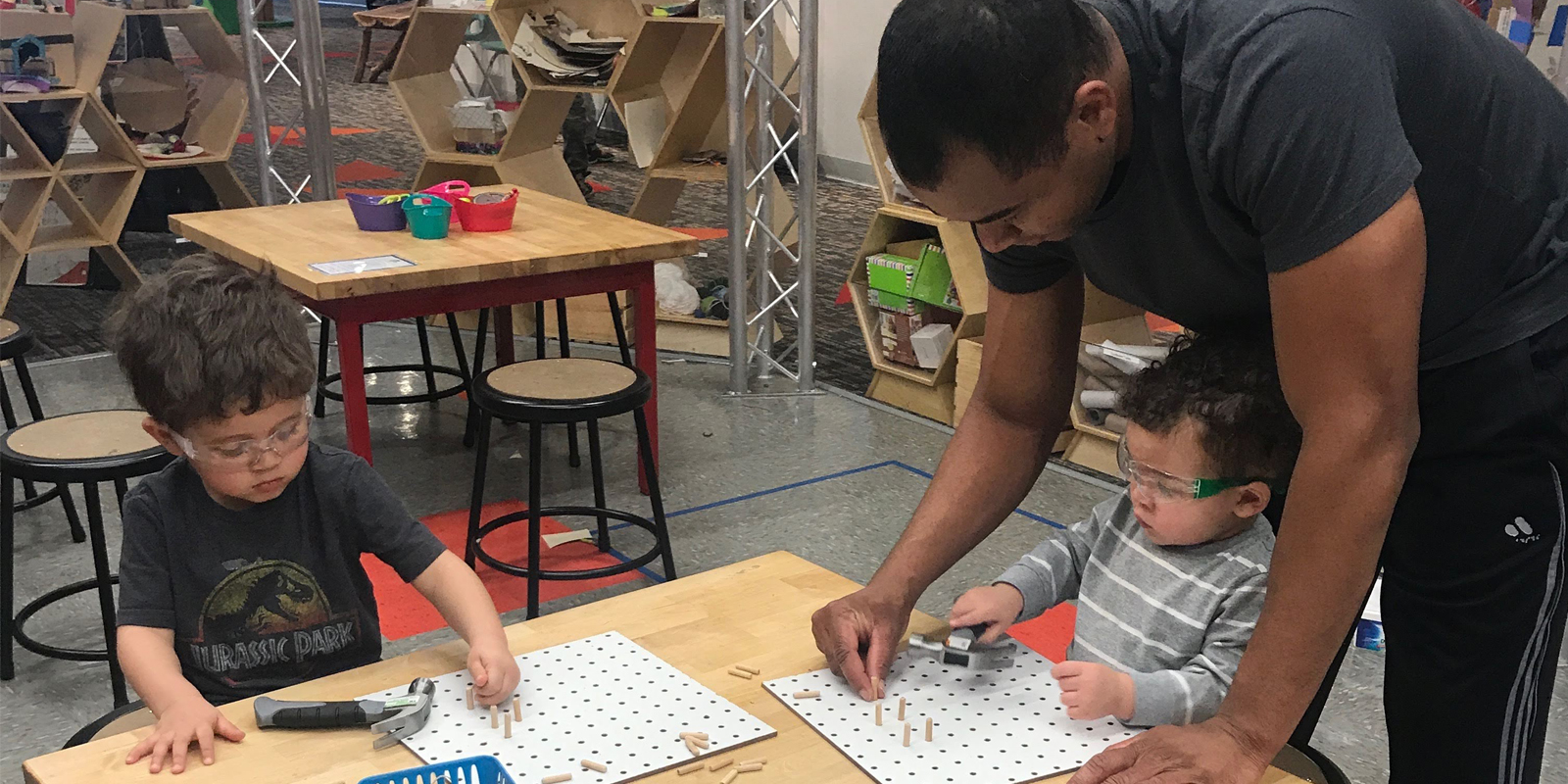 Man with dark hair with two small boys wearing goggles and playing with wooden pegboard and pegs.