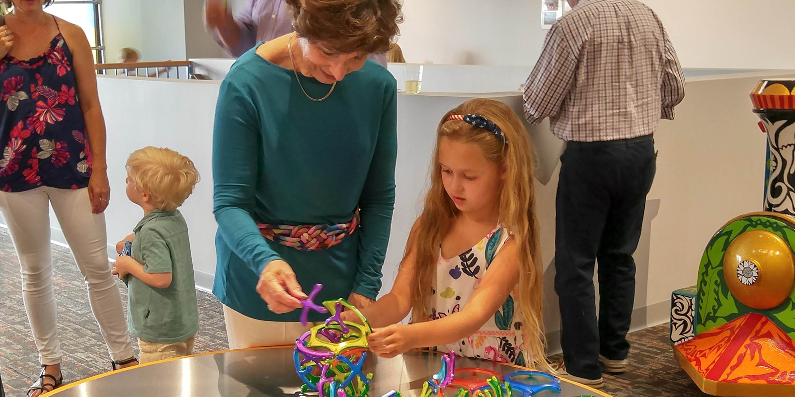 Woman in green shirt building plastic model with young blonde girl in white dress
