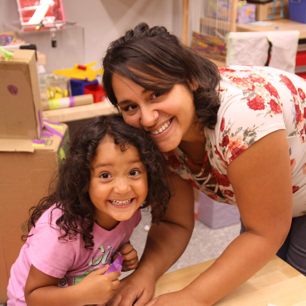 Young woman in white flowered shirt and jeans building paper crafts with young girl with dark hair and pink t-shirt.