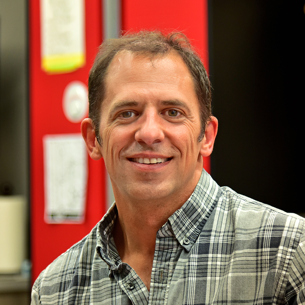 Brad Oneglia Man with brown hair wearing a black, gray and white checkered button down shirt smiling