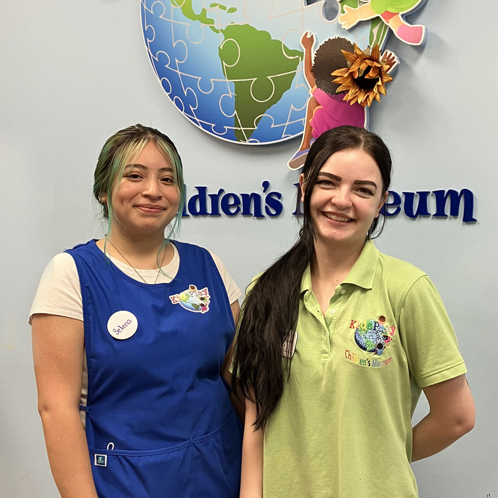 Young woman in blue KidsPlay smock stands next to young woman in KidsPlay green polo shirt in front of KidsPlay Children's Museum