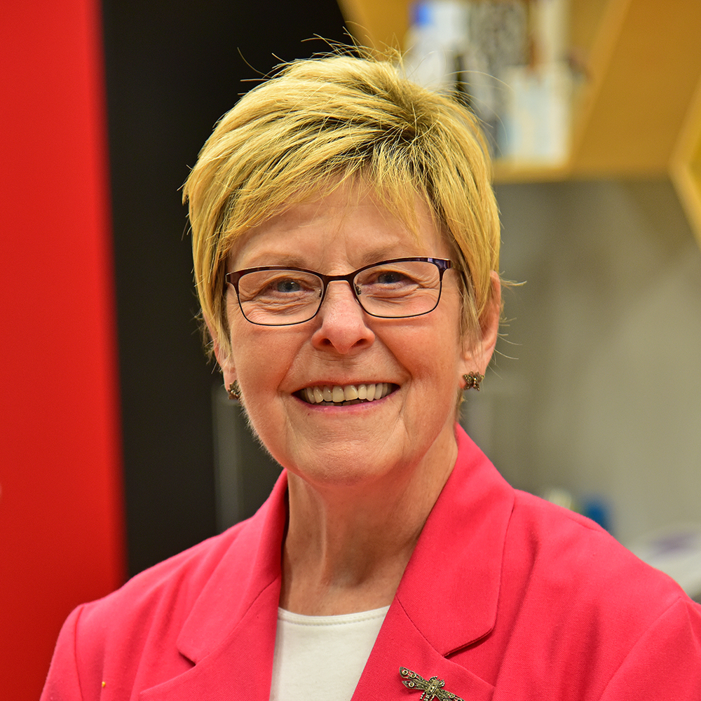 Mary Guilfoile Woman with short blonde hair and dark-rimmed glasses wearing red sports coat over a white shirt smiling.