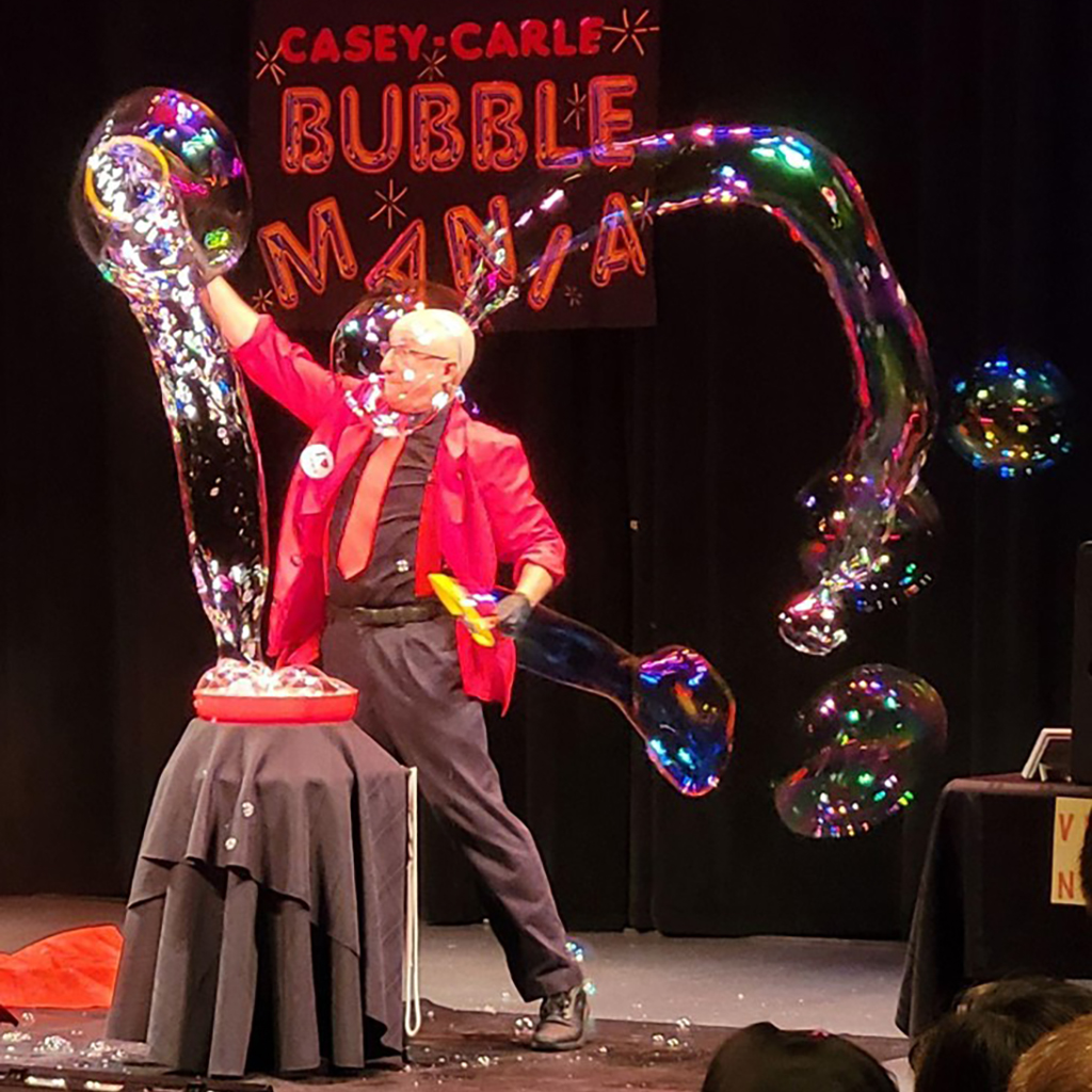 KidsPlay Birthday - Casey Carle, Bubble Mania - A guy in a magenta shirt with gray pants and shirt waving bubbles around.